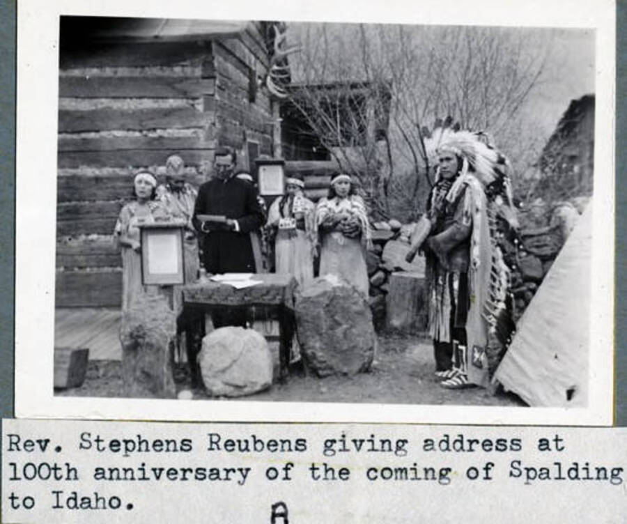 Rev. Stephens Reubens speaking, Indian in headdress in front of tepee