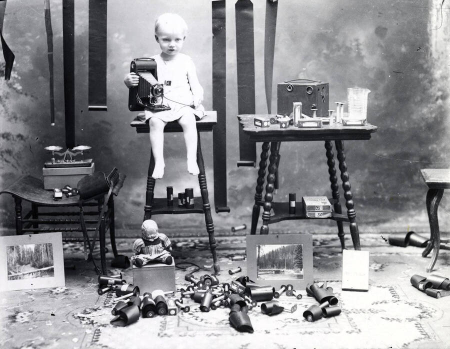 Harold, son of A.M. Whelchel, sitting on table and holding camera. Also shows film, tables, and a doll