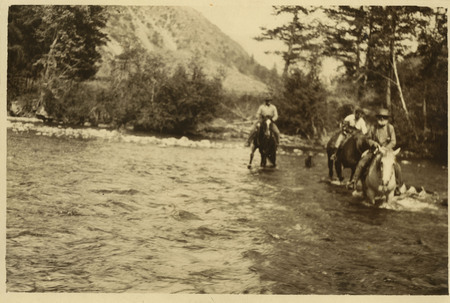 A photograph of Dave Lewis and two men fording stream on horseback. A blurry figure of a dog is at the back of the river.