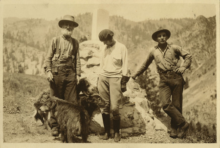 Three people and three dogs stand in front of a headstone at the Sheepeater Battlefield on Soldier Bar. Front left to right, David Lewis, Thelma Page, and an unidentifiable man.