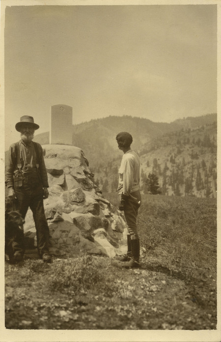 David Lewis, with two dogs at his heel, stands to the left of the headstone. Thelma Page is turned away from the camera, looking at the headstone at the Sheepeater Battlefield on Soldier Bar.