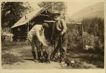 David Lewis and Thelma Page, faced away, pose in front of a log cabin. Thelma Page is bent over petting a dog, and Lewis stares at the camera while holding a gun.
