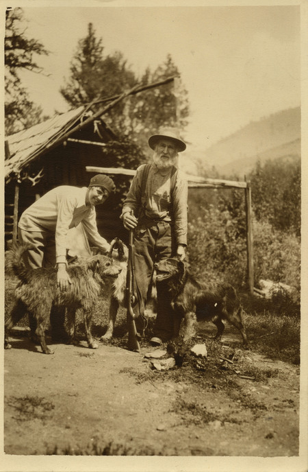 David Lewis, Thelma Page and three dogs are featured in the photograph in front of a log cabin.