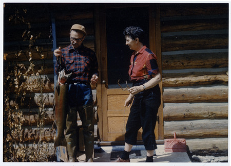 Dorothy Taylor (right) and unidentified man stare at a large trout. A door to a log cabin is in the background.