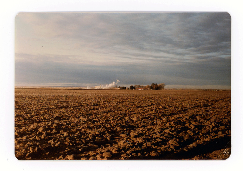 The front of a postcard decorated with a picture of an empty, tilled farm field.