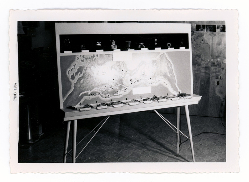 A printed picture of a top-down geographical map of an island marked with pins, displayed next to lithic artifacts.