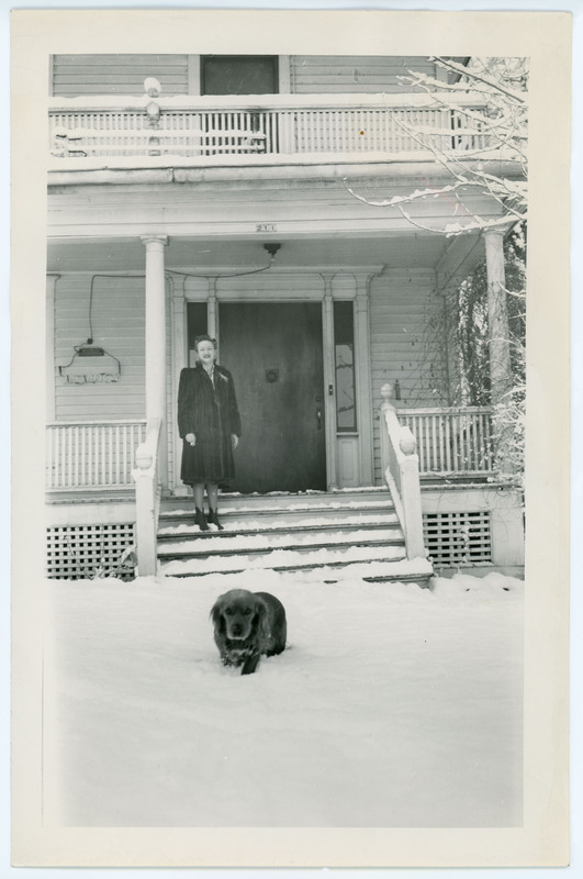 A photo of Evelyn Crabtree standing on the deck of a house. It is snowy outside and there is a dog running through the snow. On the back of the image is a note stating the house is in Twin Falls, Idaho.