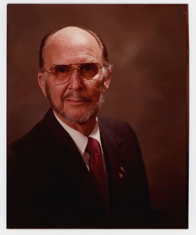 A photo of Donald Crabtree in a suit with a straight face sitting for a headshot. There are pins on his suit.