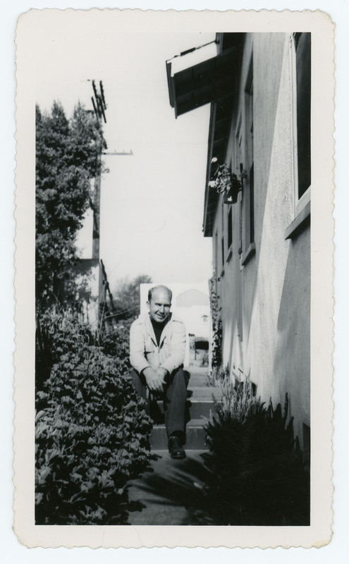 A photo of Donald Crabtree sitting on stairs outside a house. He is sitting next to foliage, and the side of the house is in the image.