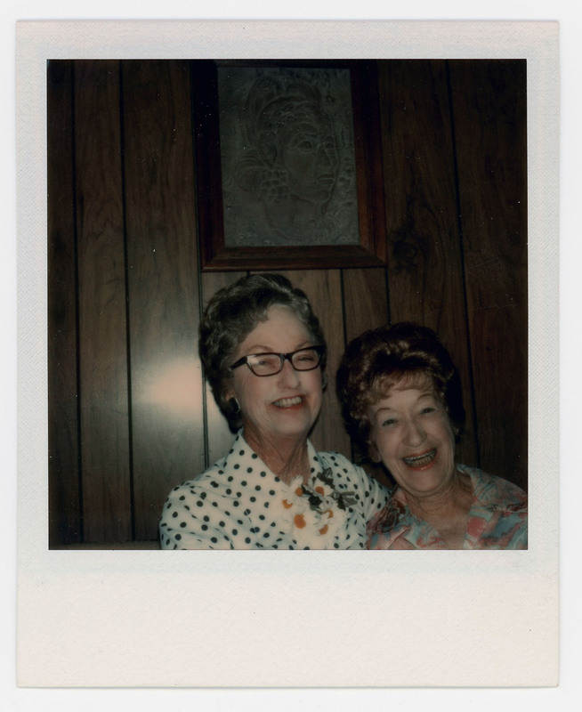 A photo of Evelyn Crabtree and another woman sitting together and laughing. There is a painting in the background on the wall.