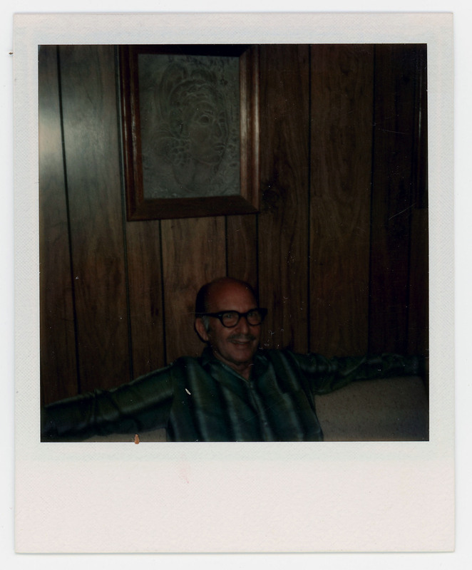 A photo of a man in glasses sitting sitting on a couch in front of a wall with a painting on it. This could be Donald Crabtree, but it is unclear as the photo is dark.