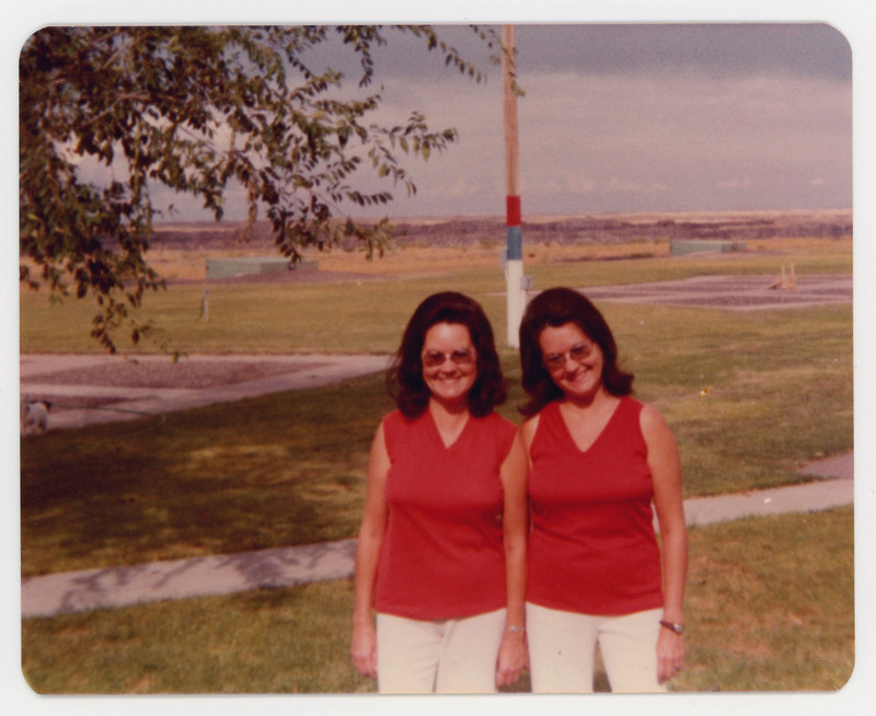 A photo of two women standing together outside in front of a field. They are wearing matching outfits and look to be twins.