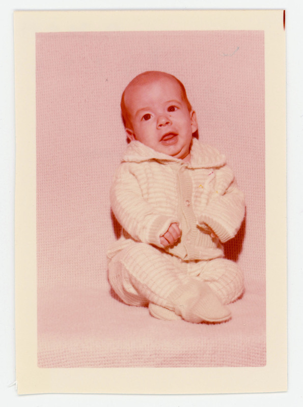 A photo of a baby sitting on a white surface. The baby is in a white striped outfit.