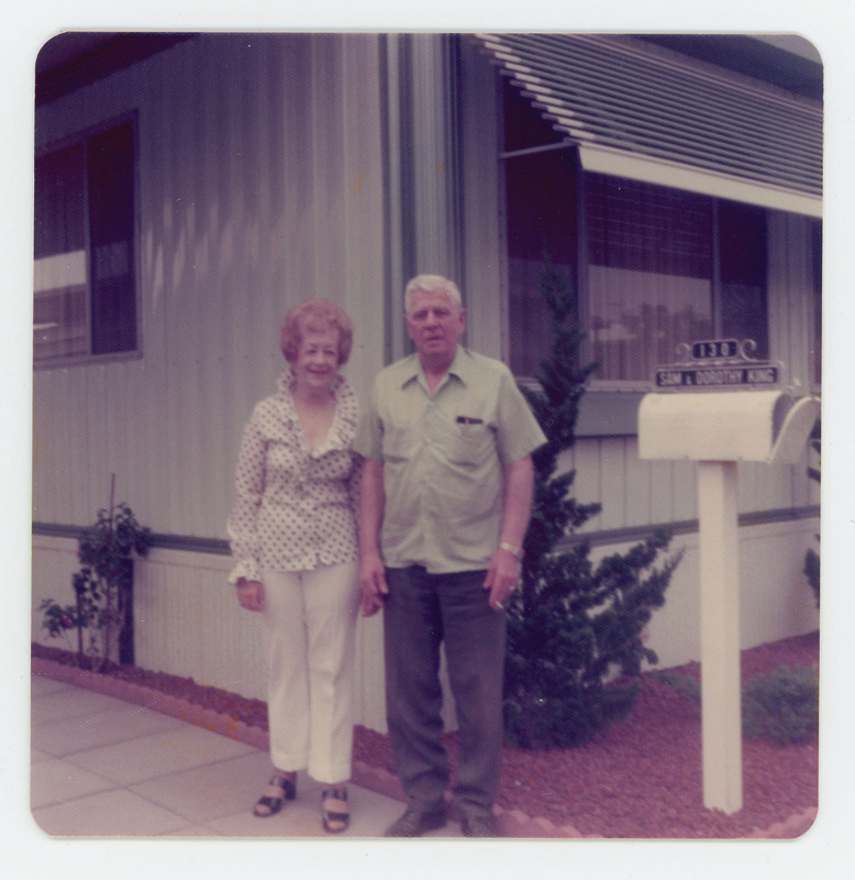 A photo of an elderly couple standing together outside a building. There is a mailbox next to them labeled Sam and Dorothy King.