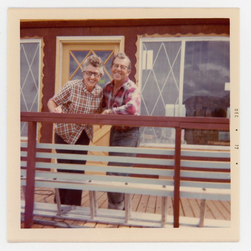 A photo of a couple standing together outside a house or building. They are on a deck and leaning on the railing.