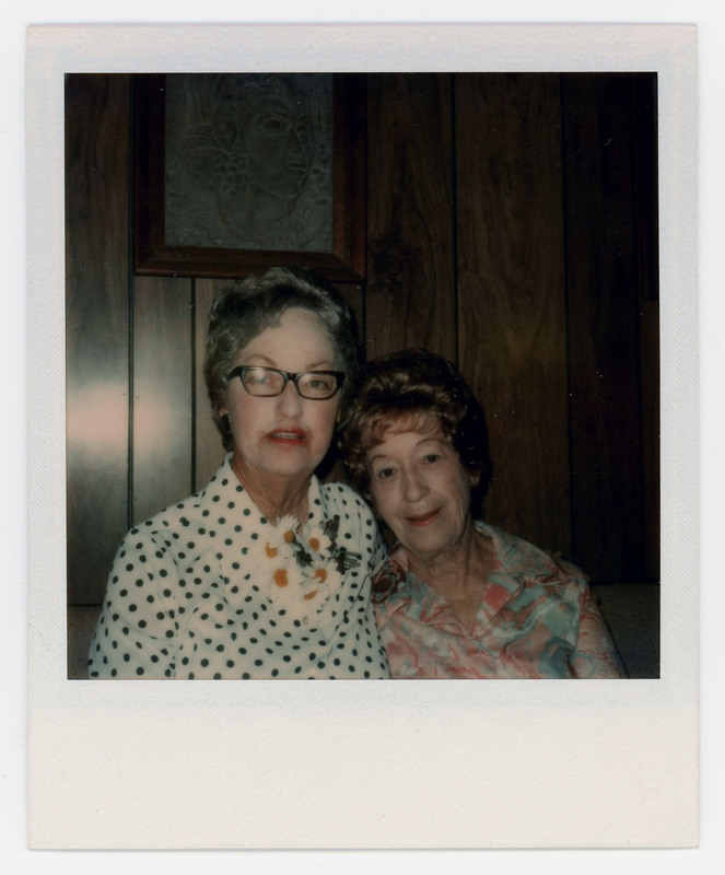 A photo of Evelyn Crabtree and another woman sitting closely together on a couch in front of a painting. Evelyn is wearing flowers on the neck of her blouse.