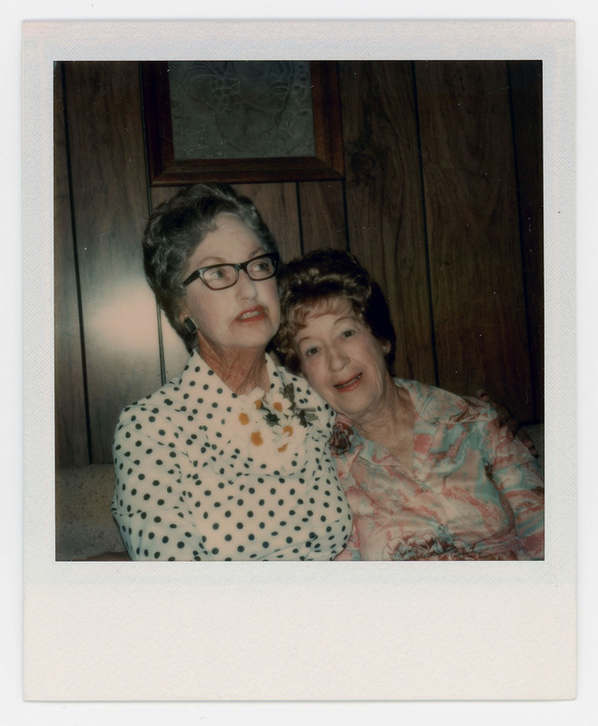 A photo of Evelyn Crabtree and a friend sitting close together on an couch. Evelyn is not looking at the camera. There is a painting on the wall behind them.