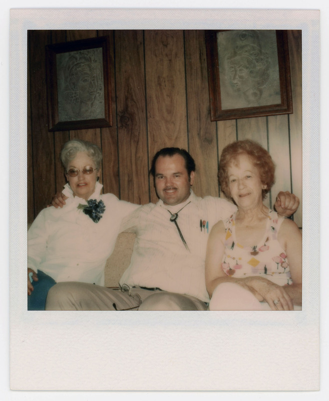 A photo of a man sitting between two women with his arms around them. They are on a couch and there are two paintings on the wall behind them.