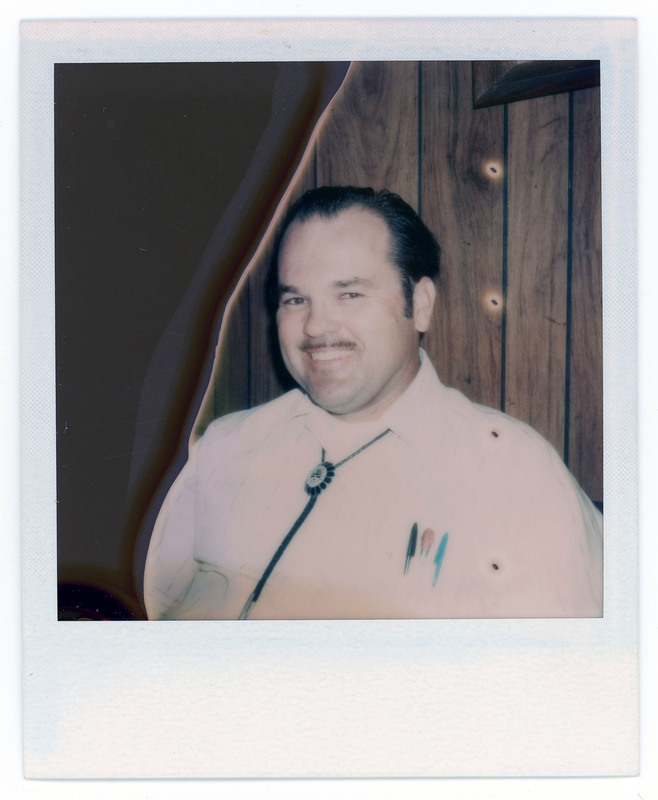 A photo of a man smiling in front of a wood wall. The man is wearing a bolo tie and has three pens in the pocket of his shirt.