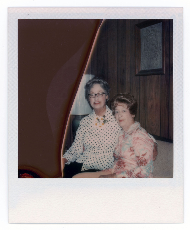 A photo of Evelyn Crabtree and a friend sitting close together on a couch. There is a lamp behind them and a painting on the wall. Evelyn has her arm around the friend.