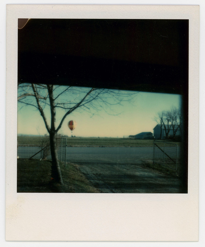 A photo of a field with an air balloon distant in the background. There is an open fence in the foreground and a house nearby.