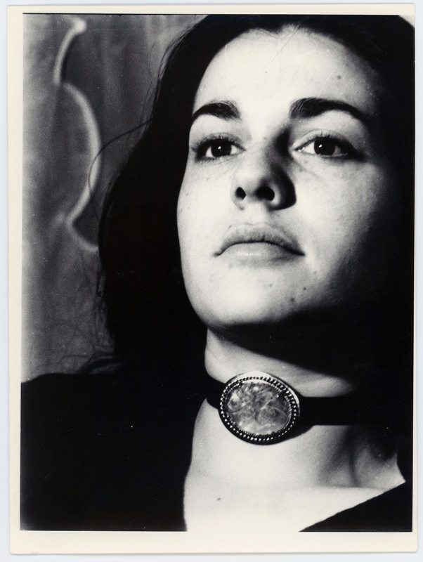 A photo of a woman with dark hair not looking directly at the camera. She is wearing a choker necklace with a large stone.