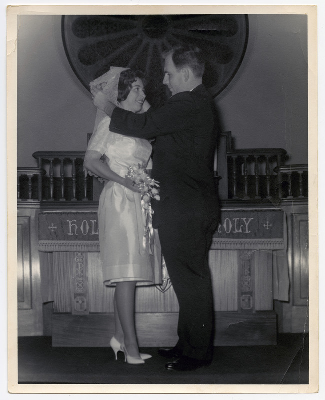 A photo of a woman in a wedding dress holding flowers and standing in front of a man. The man is pulling her veil back. They are in a church.