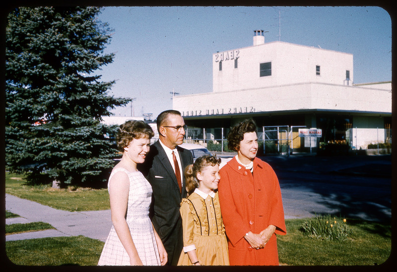 A photographic slide displaying two adults and two children standing outside of a SEARS store. From left to right there is a woman in a red coat, a young child in an orange dress, a man with glasses in a suit, and an older child in a white dress. They are facing slightly left, away from the camera.