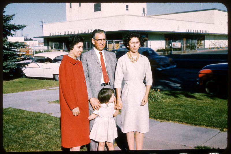 A photographic slide displaying three adults and one child standing outside of a SEARS store. From left to right there is a woman in a white dress, a man with glasses in a suit, a young girl in a white dress, and a woman in a red coat. The man and the woman on the left face the camera, while the woman on the right and the young child face left of the camera.