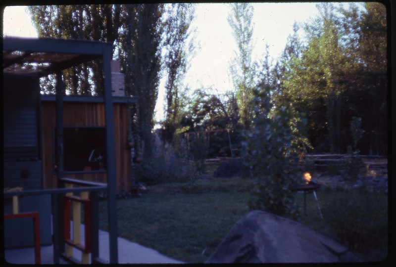 A photographic slide displaying the backyard of a house. There is a shade over the porch, a part of the house in the back, and a couple of newly planted trees in the yard.