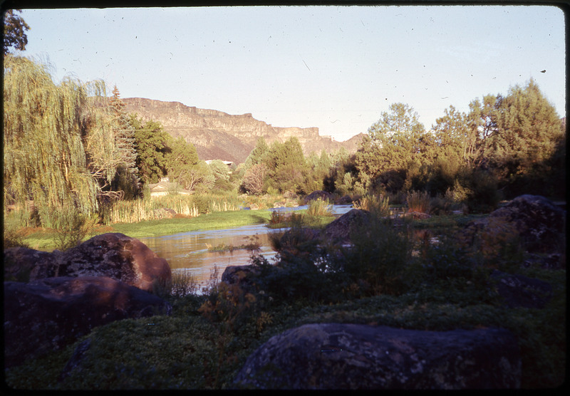 A photographic slide displaying a river in front of some foothills. The river's coast is covered in trees, save a clear areas with large rocks and small plants towards the front.