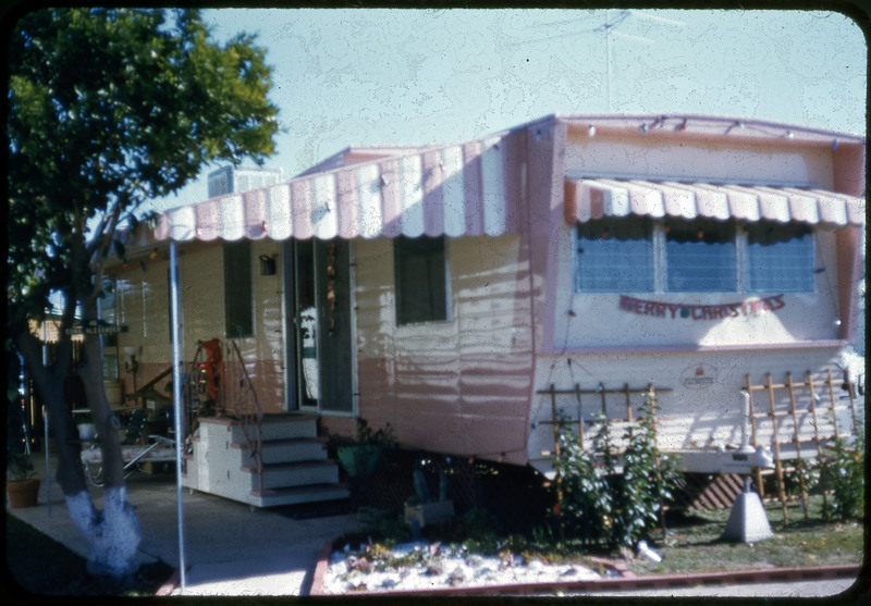 A photographic slide displaying an RV with a decorated porch area. There is a striped overhang shading the porch and two trellises with plants decorating the front. A banner hung at the front of the RV says "Merry Christmas".