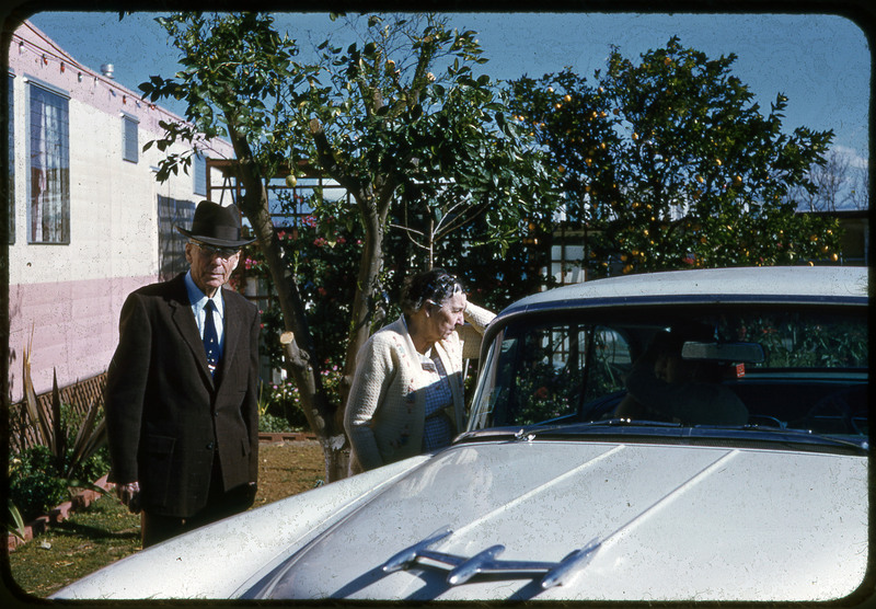 A photographic slide displaying a man and a woman behind a car. Behind them is the side of an RV and a line of small trees. The man, left, is wearing a suit and hat. The woman, right, is wearing a white jacket and looks at the car.