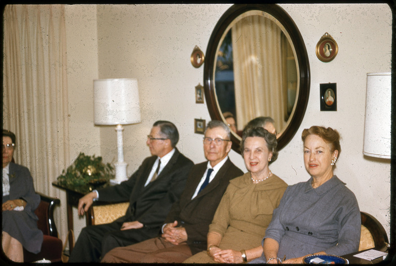 A photographic slide displaying five people sitting together inside. Two men in suits and two women in coats sit on a couch, while one woman in a jacket sits at an armchair to their left. The woman on the left is cut-off by the image. The interior is decorated with lamps, photographs, and a mirror.