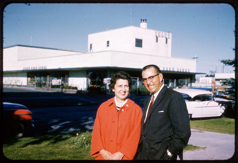 A photographic slide displaying a man and a woman standing outside a SEARS store together. The woman, left, wears a red coat while the man, right, wears glasses and a suit. They both smile and face the camera.