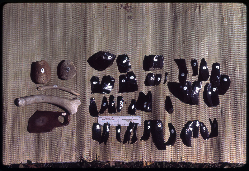 A photographic slide of a series of obsidian blades and assorted flintknapping tools accompanied by a measuring scale.