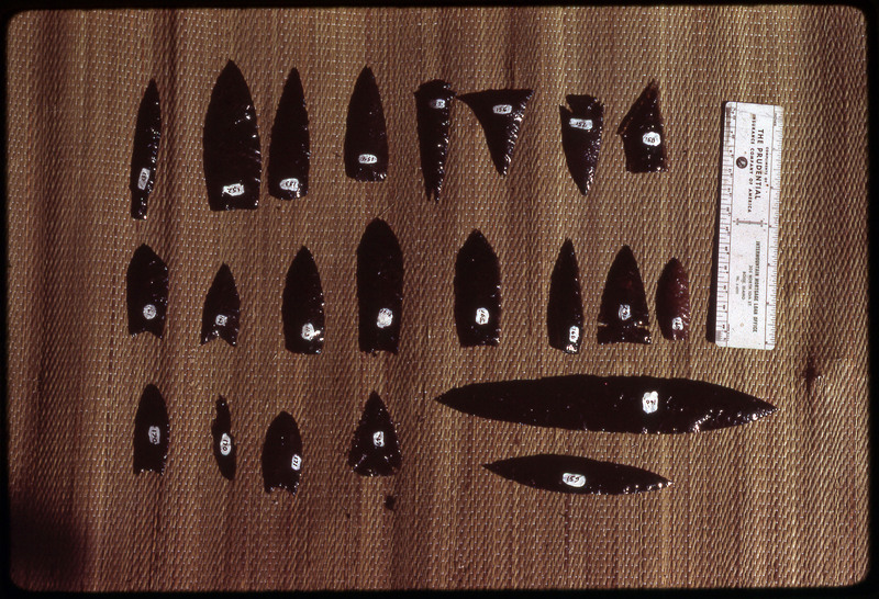 A photographic slide of a series of obsidian points and bifaces accompanied by a measuring scale.