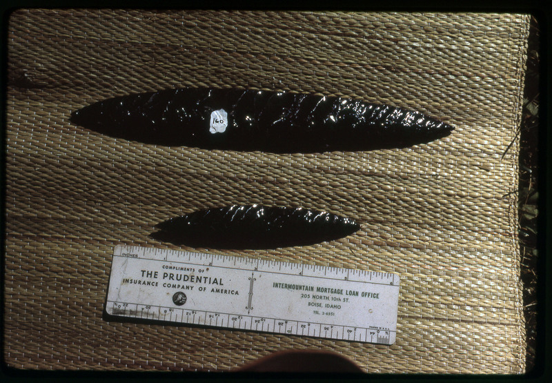 A photographic slide of two obsidian bifaces accompanied by a measuring scale. The larger one is labelled "160".