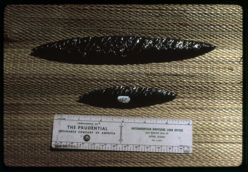 A photographic slide of a two obsidian bifaces accompanied by a measuring scale. The smaller one is labelled "159".