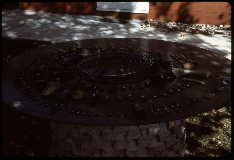 A photographic slide of a series of obsidian implements and flintknapping tools organized in concentric circles on a table.