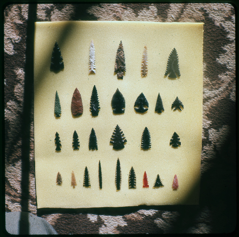 A photographic slide of a series of lithic arrowheads of various designs, materials, and sizes organized in a decorational pattern.