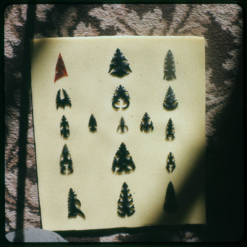 A photographic slide of a series of lithic arrowheads of various designs, materials, and sizes organized in a decorational pattern.