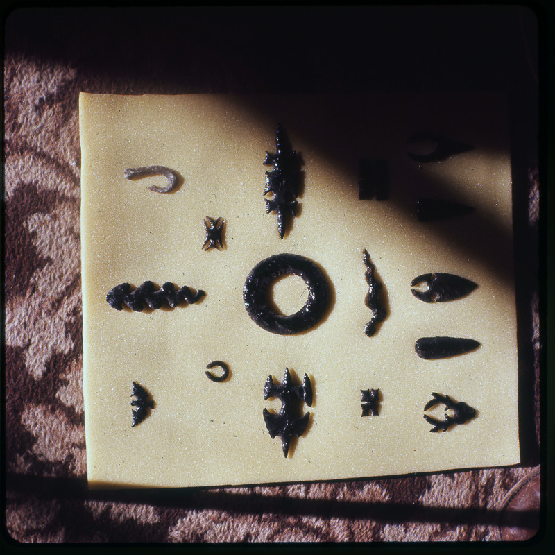 A photographic slide of a series of lithic arrowheads and unique shapes of various designs, materials, and sizes organized in a decorational pattern.