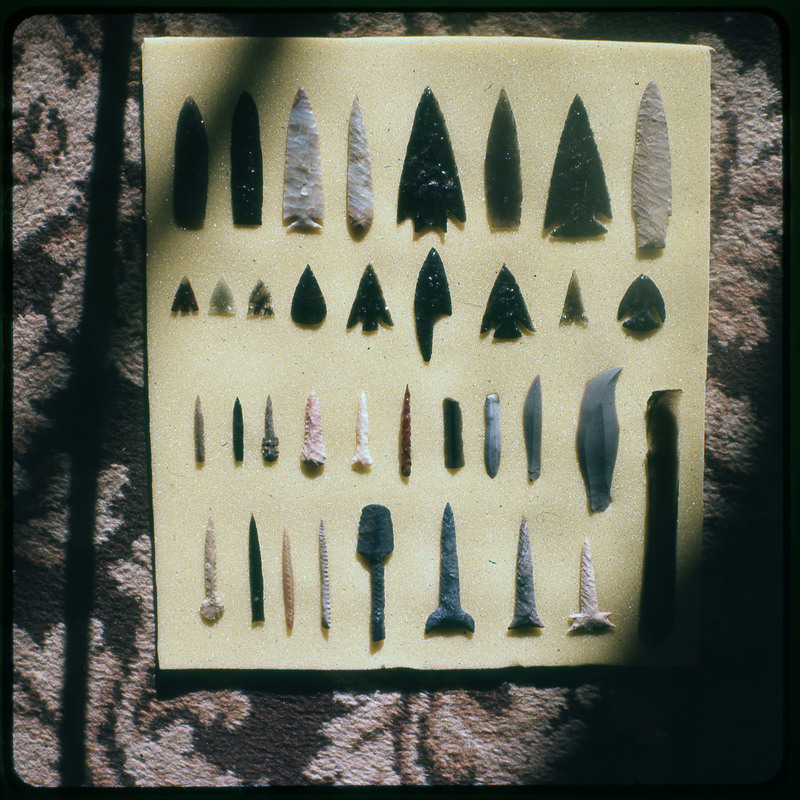 A photographic slide of a series of lithic arrowheads, blades, and punches of various designs, materials, and sizes organized in a decorational pattern.