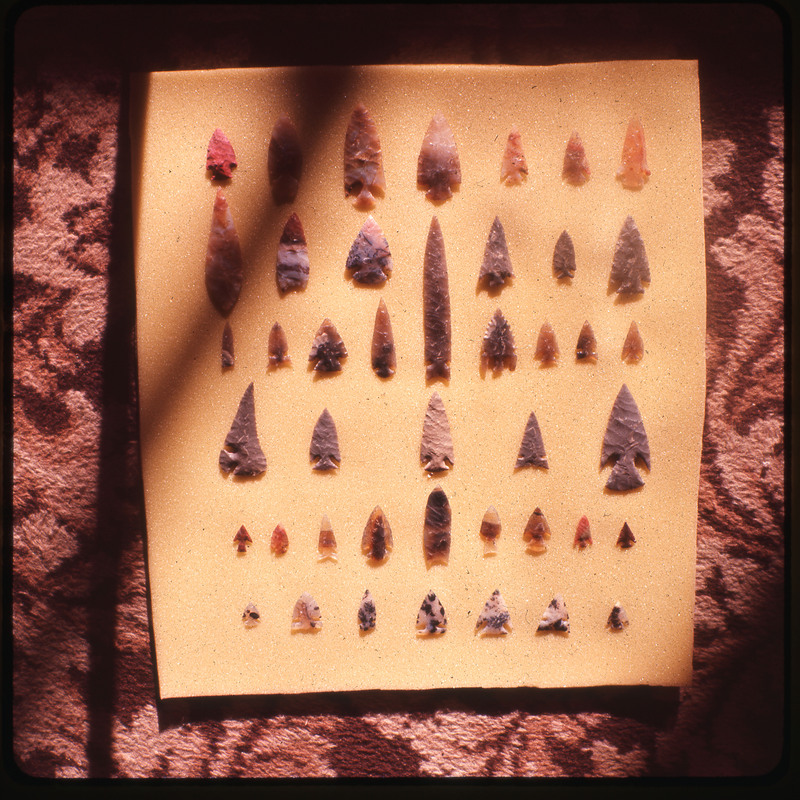 A photographic slide of a series of lithic arrowheads of various designs, materials, and sizes organized in a decorational pattern.