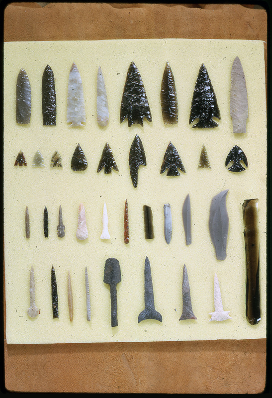 A photographic slide of a series of lithic arrowheads, blades, and punches of various designs, materials, and sizes organized in a decorational pattern.