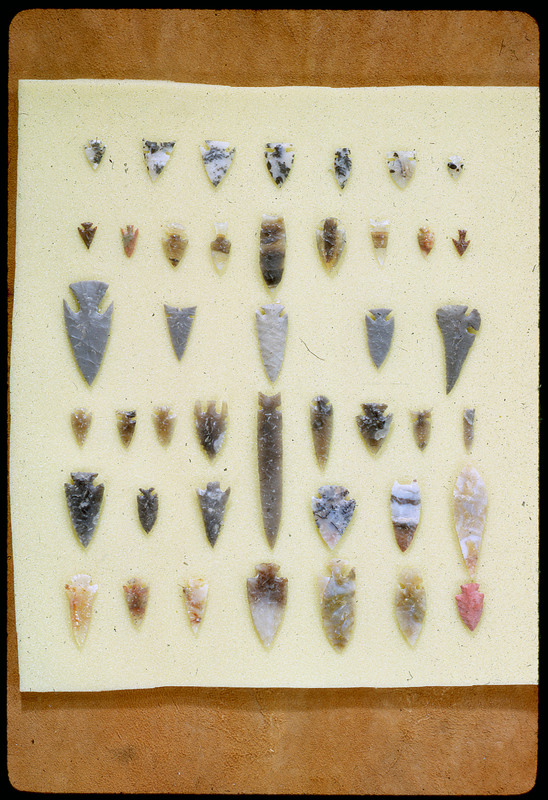 A photographic slide of a series of lithic arrowheads of various designs, materials, and sizes organized in a decorational pattern.