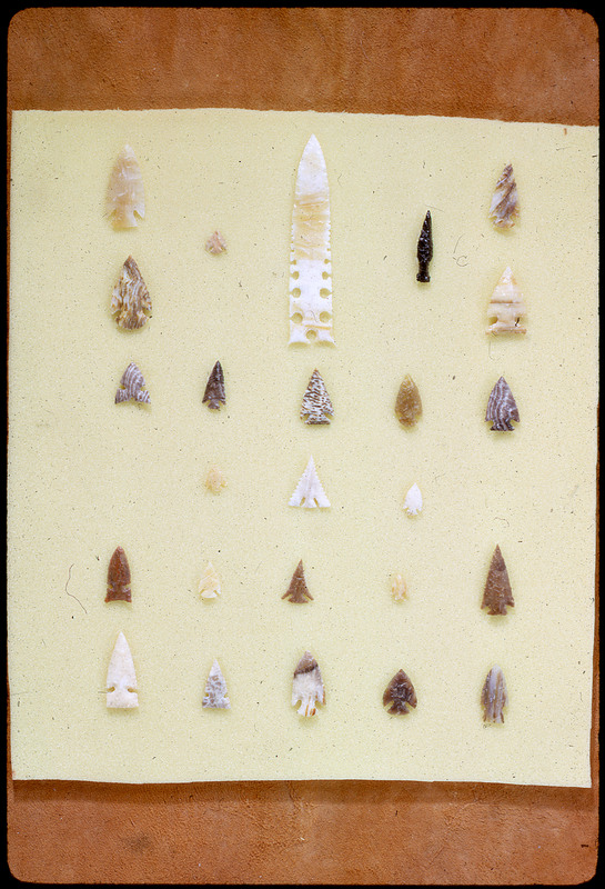 A photographic slide of a series of lithic arrowheads of various designs, materials, and sizes organized in a decorational pattern.