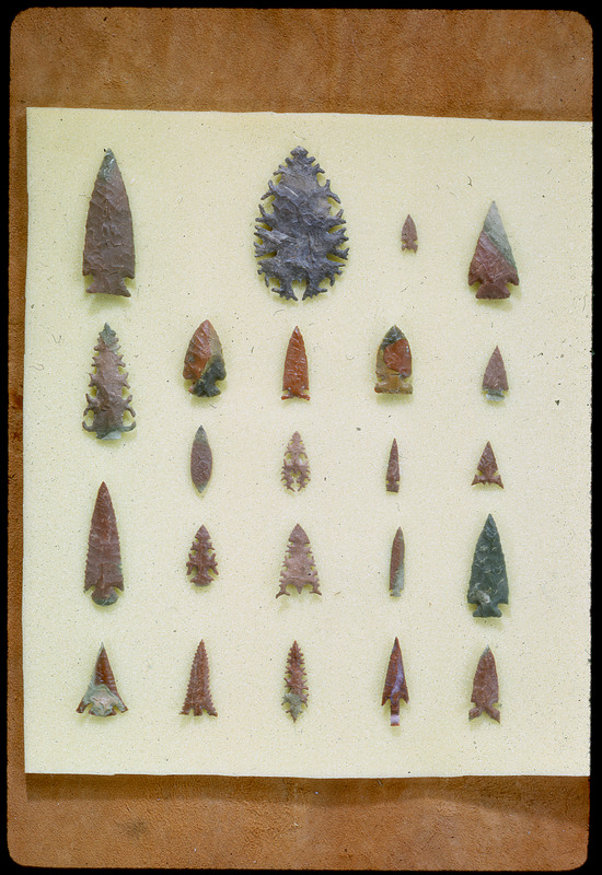 A photographic slide of a series of lithic arrowheads of various designs, materials, and sizes organized in a decorational pattern.