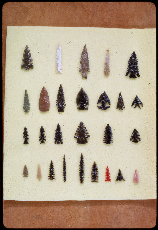 A photographic slide of a series of lithic arrowheads of various designs, materials, and sizes organized in a decorational pattern.